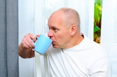 Beyaz tişörtlü, elinde mavi bardakla pencereden dışarı bakan olgun kel adam.