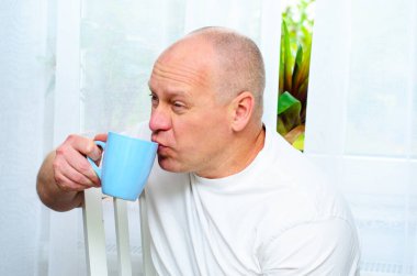 Beyaz tişörtlü, elinde mavi bardakla pencereden dışarı bakan olgun kel adam.