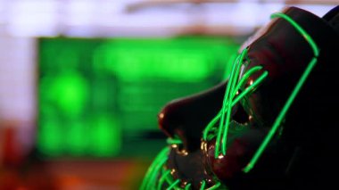 Parlayan yeşil bir hacker maskesi sinema siber masasında duruyor. Üçlü monitörler, yeşil-turuncu ışıklandırmalı karanlık bir odada şifreli kod yayıyor. Dijital sızma atmosferini çağrıştırıyor..