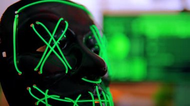 Parlayan yeşil bir hacker maskesi sinema siber masasında duruyor. Üçlü monitörler, yeşil-turuncu ışıklandırmalı karanlık bir odada şifreli kod yayıyor. Dijital sızma atmosferini çağrıştırıyor..