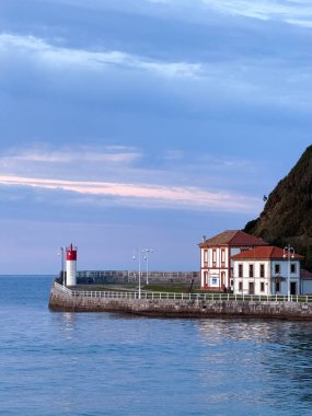 Sakin bir okyanus koyunun kıyısındaki küçük bir binanın yanında kırmızı ve beyaz bir fener duruyor..