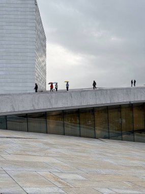 Visitors walking along the angled rooftop of the Oslo Opera House under cloudy skies.