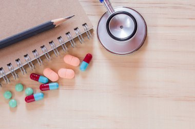 Sağlık ekipmanları - Not defteri, mavi steteskop ve çalışma odasındaki ahşap masada haplı siyah kalem. Tasarım için fotokopi alanı olan üst görünüm. İş sağlığı ve tıbbi konsept