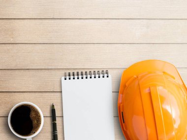 Boş not defteri, turuncu güvenlik kaskı ve kahverengi ahşap masa arkasında bir fincan kahve. Metin veya herhangi bir tasarım için kopyalama alanı olan en üstteki görünüm. Masa tahtası kağıt konsepti üzerine beyaz kağıt