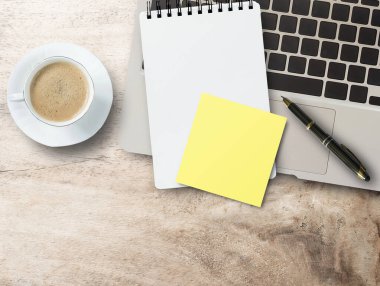 Boş sarı yapışkan kağıt, dizüstü bilgisayarda not defteri, ofis masasında kalem ve kahve fincanı. Kopyalama alanı olan üst görünüm.