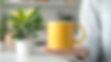 Yeşil bir bitkinin yer aldığı bulanık mutfak arka planına karşı bir elde tutulan parlak sarı bir kahve fincanı. Yaşam tarzı ve ev dekorasyonu teması için mükemmel.