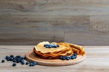 Tatlı sabah lezzeti: yaban mersinli ve şekerli ince krepler. Yaban mersini ve pudra şekeriyle süslenmiş, ahşap tabakta duran altın ince krepler. Taze pişmiş ev yemeği, geleneksel tatlı kahvaltı fikri.