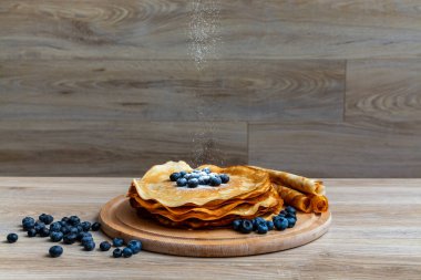 Tatlı sabah lezzeti: yaban mersinli ve şekerli ince krepler. Yaban mersini ve pudra şekeriyle süslenmiş, ahşap tabakta duran altın ince krepler. Taze pişmiş ev yemeği, geleneksel tatlı kahvaltı fikri.