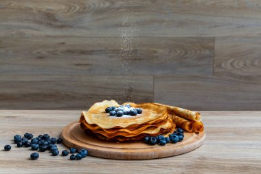 Tatlı sabah lezzeti: yaban mersinli ve şekerli ince krepler. Yaban mersini ve pudra şekeriyle süslenmiş, ahşap tabakta duran altın ince krepler. Taze pişmiş ev yemeği, geleneksel tatlı kahvaltı fikri.