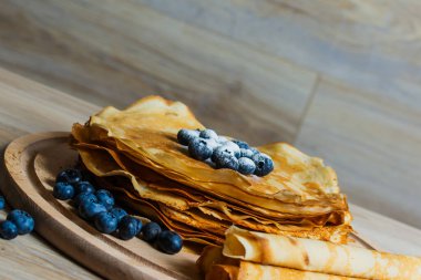 Tatlı sabah lezzeti: yaban mersinli ve şekerli ince krepler. Yaban mersini ve pudra şekeriyle süslenmiş, ahşap tabakta duran altın ince krepler. Taze pişmiş ev yemeği, geleneksel tatlı kahvaltı fikri.