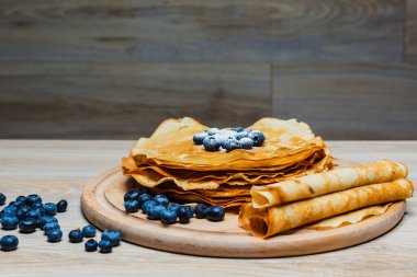 Tatlı sabah lezzeti: yaban mersinli ve şekerli ince krepler. Yaban mersini ve pudra şekeriyle süslenmiş, ahşap tabakta duran altın ince krepler. Taze pişmiş ev yemeği, geleneksel tatlı kahvaltı fikri.