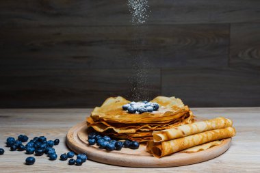 Tatlı sabah lezzeti: yaban mersinli ve şekerli ince krepler. Yaban mersini ve pudra şekeriyle süslenmiş, ahşap tabakta duran altın ince krepler. Taze pişmiş ev yemeği, geleneksel tatlı kahvaltı fikri.