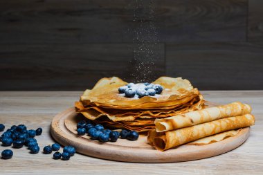 Tatlı sabah lezzeti: yaban mersinli ve şekerli ince krepler. Yaban mersini ve pudra şekeriyle süslenmiş, ahşap tabakta duran altın ince krepler. Taze pişmiş ev yemeği, geleneksel tatlı kahvaltı fikri.