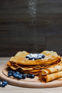 Tatlı sabah lezzeti: yaban mersinli ve şekerli ince krepler. Yaban mersini ve pudra şekeriyle süslenmiş, ahşap tabakta duran altın ince krepler. Taze pişmiş ev yemeği, geleneksel tatlı kahvaltı fikri.