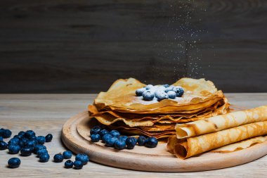 Tatlı sabah lezzeti: yaban mersinli ve şekerli ince krepler. Yaban mersini ve pudra şekeriyle süslenmiş, ahşap tabakta duran altın ince krepler. Taze pişmiş ev yemeği, geleneksel tatlı kahvaltı fikri.