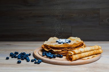 Tatlı sabah lezzeti: yaban mersinli ve şekerli ince krepler. Yaban mersini ve pudra şekeriyle süslenmiş, ahşap tabakta duran altın ince krepler. Taze pişmiş ev yemeği, geleneksel tatlı kahvaltı fikri.