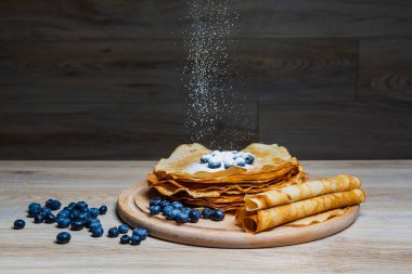 Tatlı sabah lezzeti: yaban mersinli ve şekerli ince krepler. Yaban mersini ve pudra şekeriyle süslenmiş, ahşap tabakta duran altın ince krepler. Taze pişmiş ev yemeği, geleneksel tatlı kahvaltı fikri.