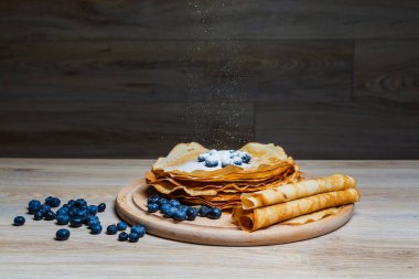 Tatlı sabah lezzeti: yaban mersinli ve şekerli ince krepler. Yaban mersini ve pudra şekeriyle süslenmiş, ahşap tabakta duran altın ince krepler. Taze pişmiş ev yemeği, geleneksel tatlı kahvaltı fikri.