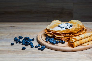 Tatlı sabah lezzeti: yaban mersinli ve şekerli ince krepler. Yaban mersini ve pudra şekeriyle süslenmiş, ahşap tabakta duran altın ince krepler. Taze pişmiş ev yemeği, geleneksel tatlı kahvaltı fikri.