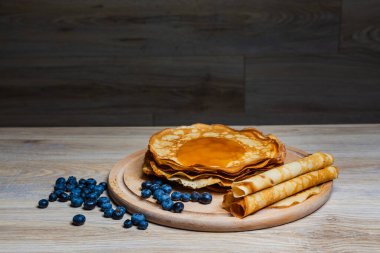 Tatlı sabah lezzeti: yaban mersinli ve şekerli ince krepler. Yaban mersini ve pudra şekeriyle süslenmiş, ahşap tabakta duran altın ince krepler. Taze pişmiş ev yemeği, geleneksel tatlı kahvaltı fikri.