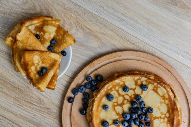 Tatlı sabah lezzeti: yaban mersinli ve şekerli ince krepler. Yaban mersini ve pudra şekeriyle süslenmiş, ahşap tabakta duran altın ince krepler. Taze pişmiş ev yemeği, geleneksel tatlı kahvaltı fikri.