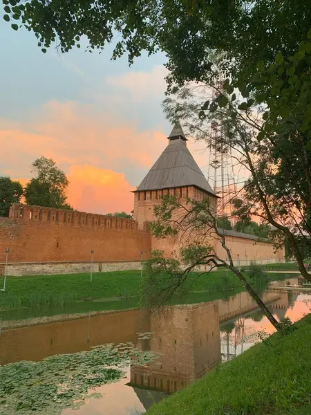 Günbatımında sakin bir hendeğe yansıyan tarihi tuğladan kale, yemyeşil ve ağaçlarla çevrili, arka planda dramatik bir gökyüzü var. Smolensk Kalesi