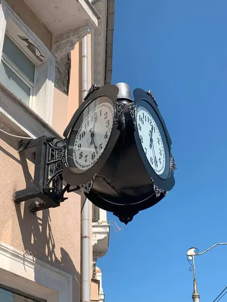 Açık mavi gökyüzünün altındaki bir binanın ön cephesine monte edilmiş klasik bir çift yüzlü saat..