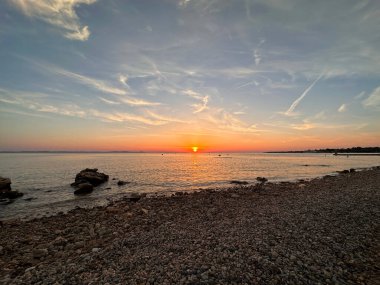 Sakin bir deniz ve dağınık bulutlarla kayalık bir sahilde sakin bir günbatımı..