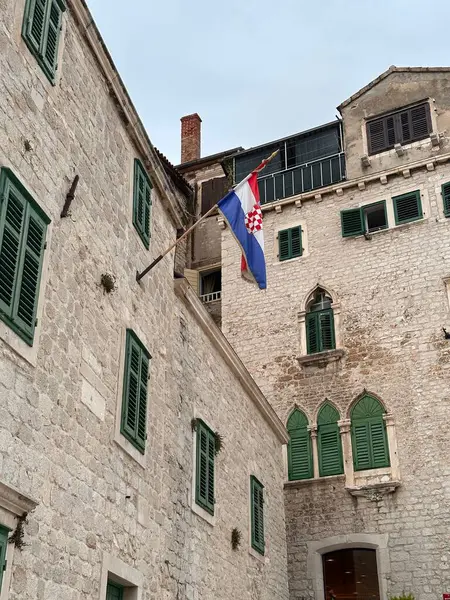 Eski bir Avrupa kentinde yeşil panjurlu tarihi taş binalar ve bir Hırvat bayrağı.