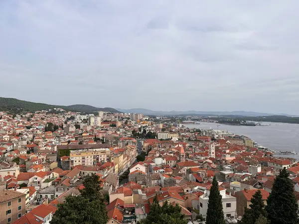 Tepeler ve bulutlu bir gökyüzünün altında sakin bir denizle çevrili, kırmızı çatılı binaları olan kıyı şehrinin havadan görünüşü..