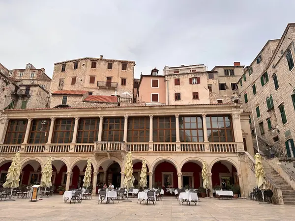 Eski taş binaları ve kemerleri olan tarihi bir Avrupa meydanı. Masa ve sandalyelerle dışarıda oturmak bir kafe ya da restoran ortamını işaret ediyor. Gökyüzü kapalı..