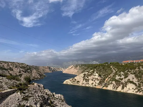 Parçalı bulutlu bir gökyüzünün altında bir köprüyle kayalık bir kanyondan akan bir nehrin manzarası..
