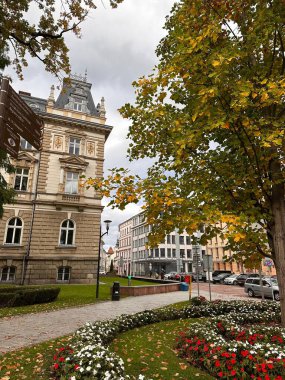 Süslü mimarisi olan tarihi bir Avrupa binası sonbahar yapraklı bir ağacın yanında duruyor. Sahnede renkli çiçekleri ve kaldırım taşı yolu olan manikürlü bir bahçe var..