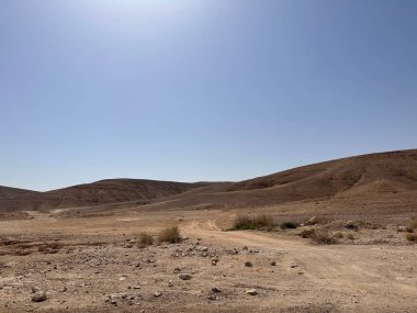 Açık mavi gökyüzünün altında yuvarlanan tepeleri olan çorak bir çöl manzarası.