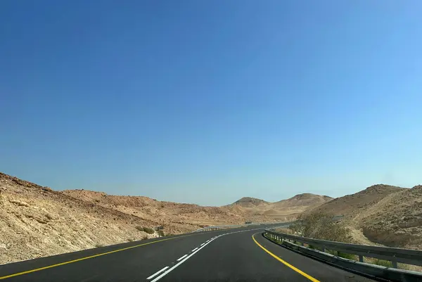 Manzaralı bir çöl otoyolu, berrak mavi gökyüzünün altında kurak, kayalık tepeler boyunca kıvrılır..