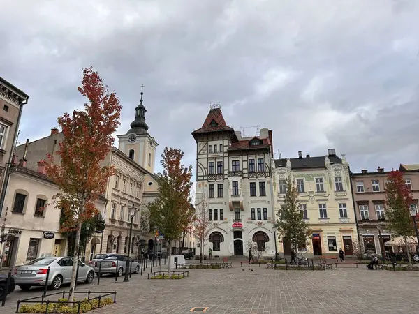 Çeşitli mimari tarzlara sahip tarihi binaların yer aldığı pitoresk bir kasaba meydanı. Kırmızı yapraklı sonbahar ağaçları meydana dizilir ve bulutlu bir gökyüzü karamsar bir atmosfer ekler..