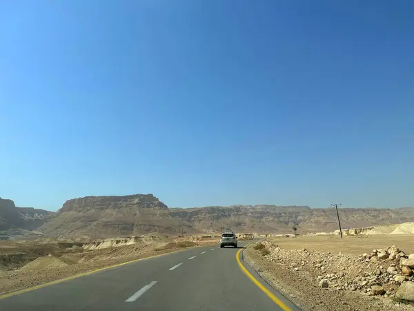 Bir araba, açık mavi bir gökyüzünün altında, arka planda dağlar olan bir çöl manzarasında dolambaçlı bir yolda seyahat eder..