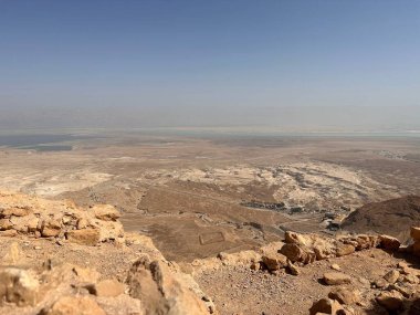 Uçsuz bucaksız bir çöl manzarasının panoramik görüntüsü kayalık arazi ve açık mavi gökyüzünün altındaki uzak dağları gösteriyor. Sahne, seyrek bitki örtüsü ve dinginlik duygusuyla bölgenin kurak güzelliğini yakalıyor..