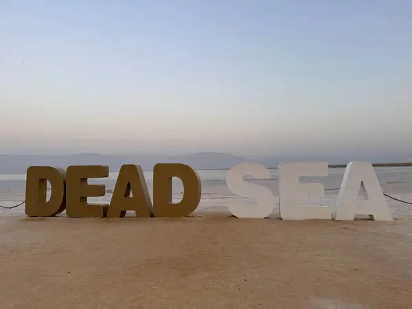 Büyük 3D harflerle yazılan 'Ölü Deniz' kumlu bir sahilde sakin bir deniz ve arka planda dağlar.