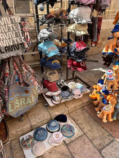Çeşitli şapka, çanta ve hediyelik eşyalar içeren canlı bir piyasa sahnesi. Sergide 'İsrail' damgalı kapaklar, renkli çantalar ve taş bir yüzey üzerindeki dekoratif eşyalar yer alıyor..