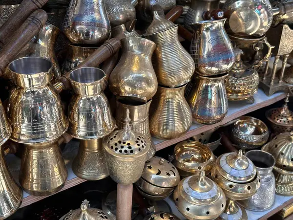 Piyasa tezgahında sergilenen süslü pirinç kahve demlikleri ve geleneksel metal yazılımlardan oluşan bir koleksiyon. Karmaşık tasarımlar ve zanaatkarlık sergiliyor..