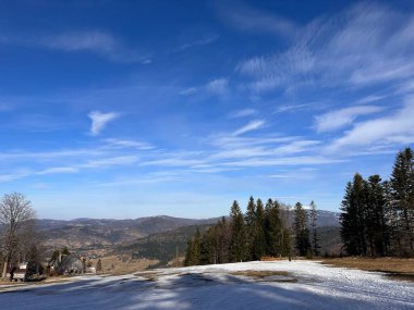 Kış manzarası, karlı bir ön plan, arka planda yuvarlanan tepeler ve ince bulutlu açık mavi bir gökyüzü. Çam ağaçları sahnenin kenarına dizilir ve sakin bir atmosfer yaratır..