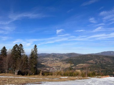 Berrak mavi gökyüzünün altındaki dağlık bir manzaranın manzarası. Ön planda kar parçaları olan çimenli bir alan yer alırken, arka planda kayan tepeler ve vadide yuva yapmış küçük bir köy yer alıyor..