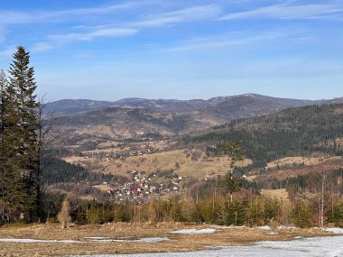 Yuvarlanan tepelerin manzarası ve açık mavi gökyüzünün altında ağaçlar ve dağlarla çevrili bir vadide yuva yapmış küçük bir köy. Yerde kar parçaları var, bu da kışı gösteriyor..