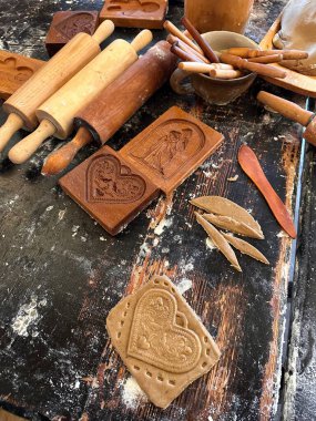 Geleneksel pişirme aletleri ve zencefilli kek kalıplarının yer aldığı kırsal bir mutfak sahnesi. Yuvarlanan raptiyeler, ahşap çatal bıçaklar ve kalp şeklindeki kurabiyeler, sıcak, nostaljik bir atmosferi çağrıştıran yıpranmış ahşap bir masaya saçılmış durumda..