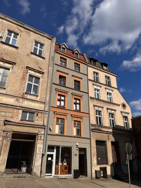 Gri ve turuncu modern bir cepheye sahip mimari tarzların karışımına sahip tarihi bir bina. Binanın birden fazla katı var, pencereleri ve zemin katında bir dükkan girişi var. Gökyüzü parçalı bulutlu.