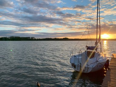 Sakin bir gölün üzerinde sakin bir günbatımı, ahşap bir iskeleye yanaşmış bir yelkenli. Gökyüzü turuncu ve sarı ılık renkleri yansıtan bulutlarla dolu. Huzurlu bir atmosfer yaratıyor..