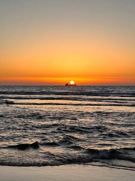 Okyanusun üzerinde sakin bir günbatımı, ufukta güneş batıyor ve ön planda bir tekne silueti var. Gökyüzü sıcak turuncu ve sarıya boyanmıştır. Suyun yüzeyine yansımaktadır..