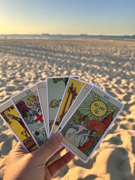 Gün batımında bir plaj geçmişine karşı tarot kartları tutan bir el. Kartlarda güneş ve diğer sembolik betimlemeler de dahil olmak üzere çeşitli çizimler yer alıyor. Kumlu sahil ve sakin deniz sakin bir atmosfer yaratır..