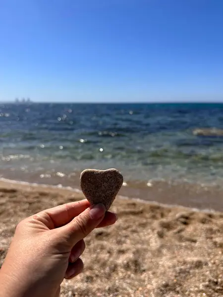 Sakin bir plaj zeminine karşı kalp şeklinde bir taşı tutan bir el. Okyanus berrak mavi gökyüzünün altında parıldıyor, sakin ve romantik bir atmosfer yaratıyor..